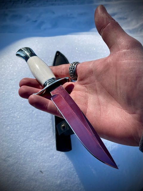 Мужская рука держит эксклюзивную финку с ярко-красным клинком и рукоятью из белого рога над сверкающим снегом.