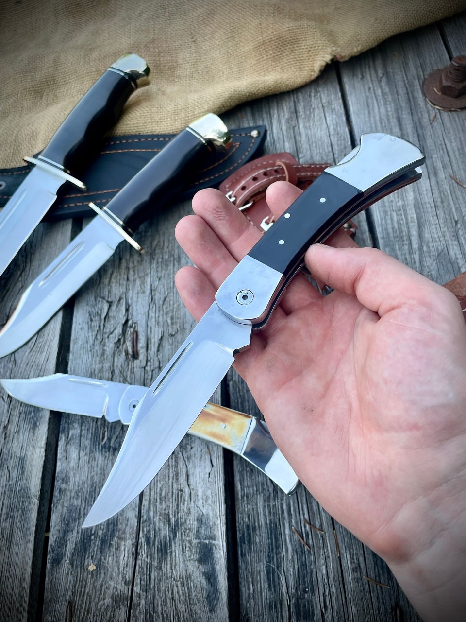 Раскрытый складной нож Buck 110 с рукоятью из чёрного G10 в руке, рядом на столе — другие ножи и нож с рукоятью из рога лося
