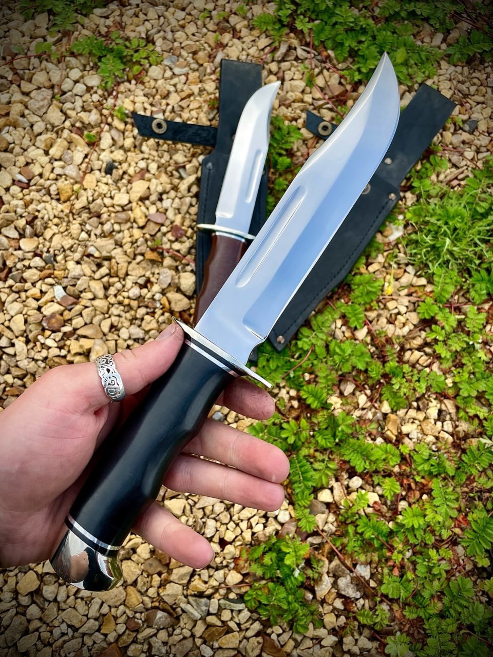 Нож Buck 120 General с чёрной рукоятью в руке крупным планом, за ним второй нож с коричневой рукоятью; клинки из стали D2 с долом на фоне щебня и ножен