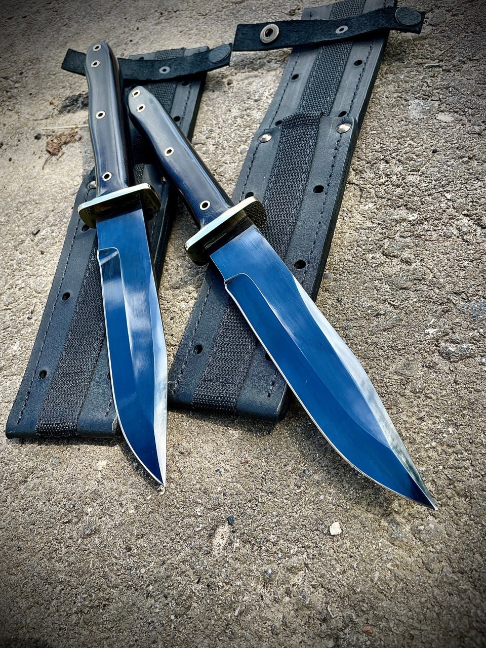 Два ножа Warrior Bowie Black Series в полностью черном тактическом исполнении лежат на бетоне рядом с ножнами.