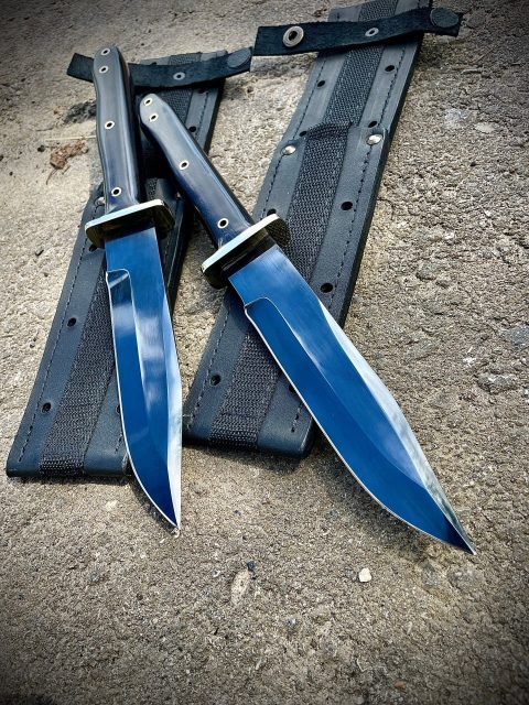 Два ножа Warrior Bowie Black Series в полностью черном тактическом исполнении лежат на бетоне рядом с ножнами.