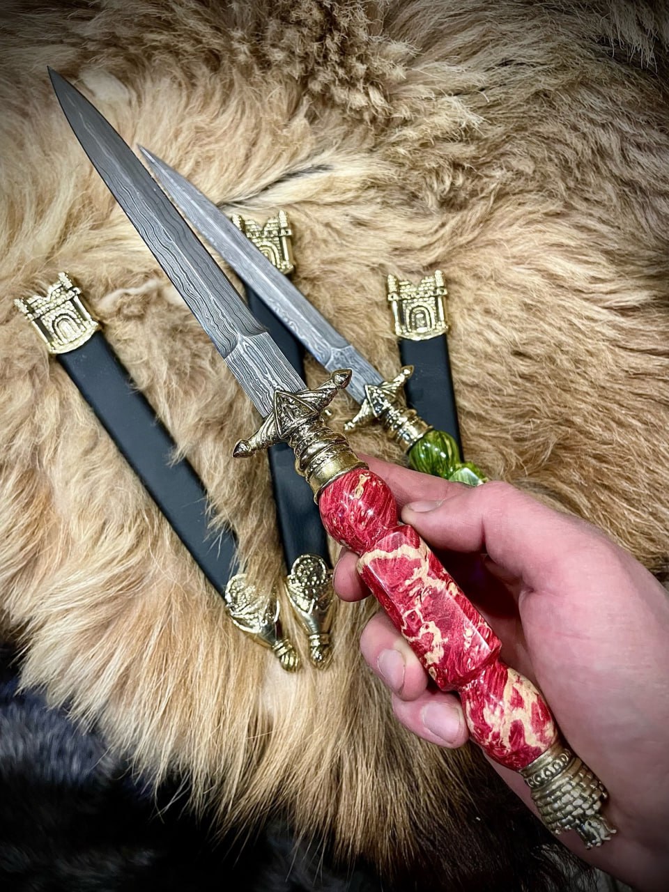 Кинжал «Донжон» с рукоятью из красной мраморовидной смолы в руке на фоне овечьей шкуры, рядом лежат экземпляры с синей и зелёной рукоятью и кожаные ножны