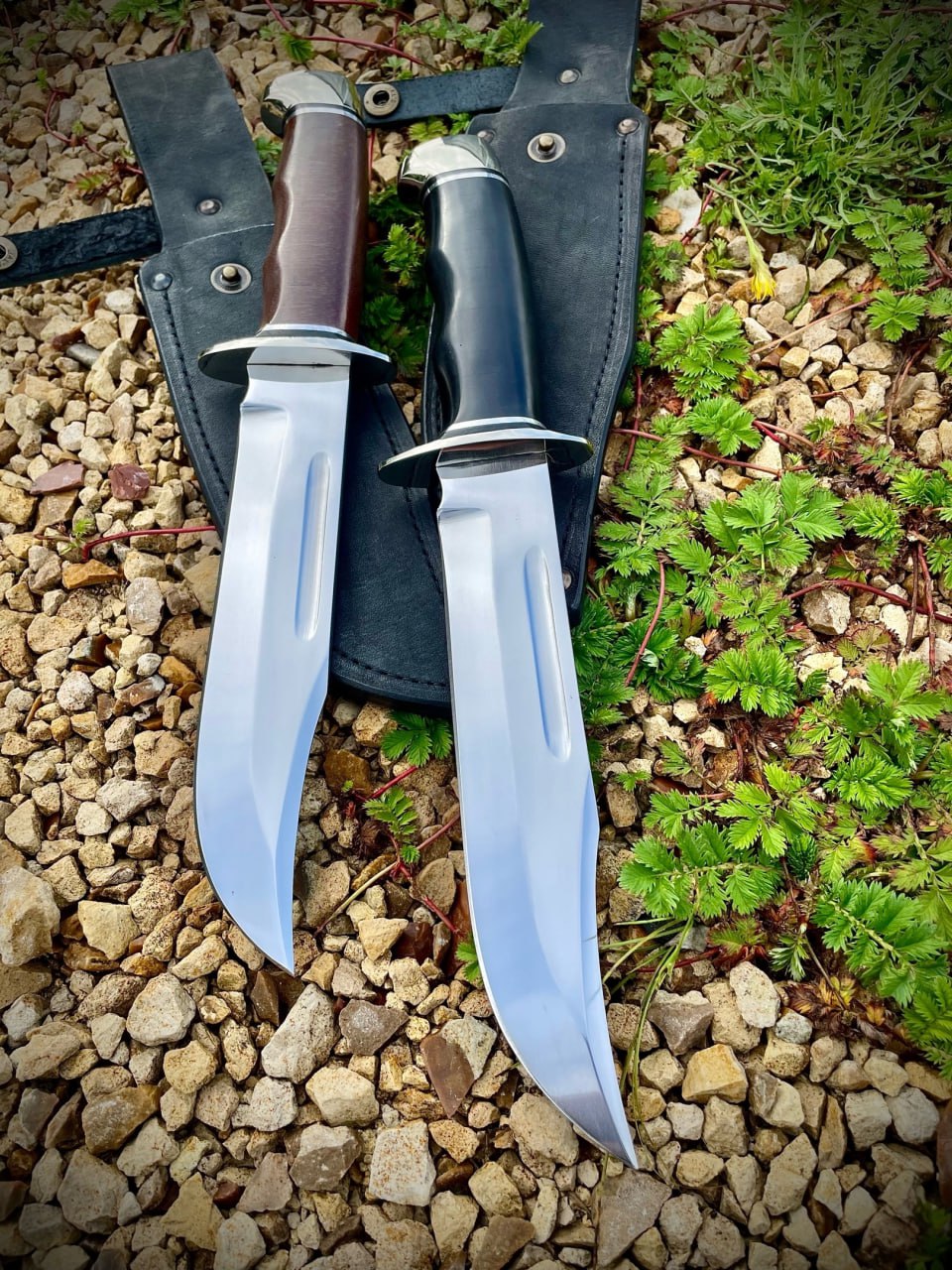 Два ножа Buck 120 General стоят рукоятями вверх на фоне щебня и зелени: коричневая текстолитовая и чёрная рукояти с полированными навершиями, большие клинки с долом
