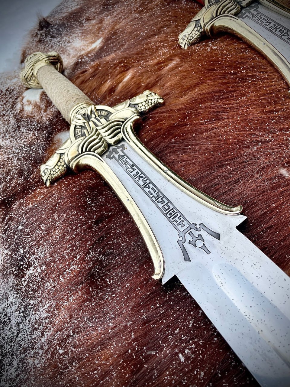 Крупный план гарды и клинка меча Конана с рунической гравировкой и литыми головами змей на фоне шкуры, рядом виден второй меч
