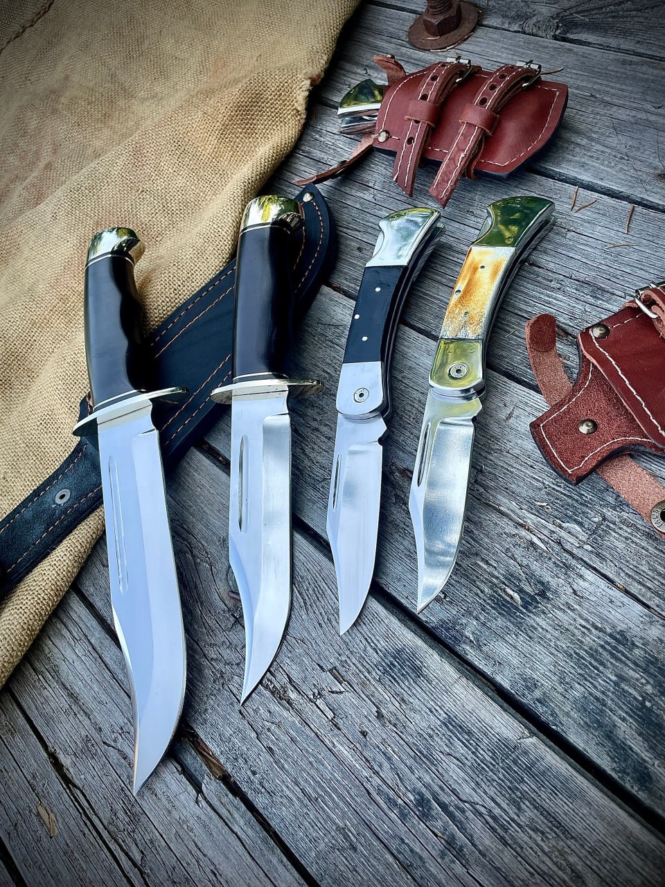 Складные ножи Buck 110 с рукоятями из чёрного G10 и рога лося рядом с охотничьими ножами с чёрными рукоятями на деревянном столе, вокруг — кожаные ножны