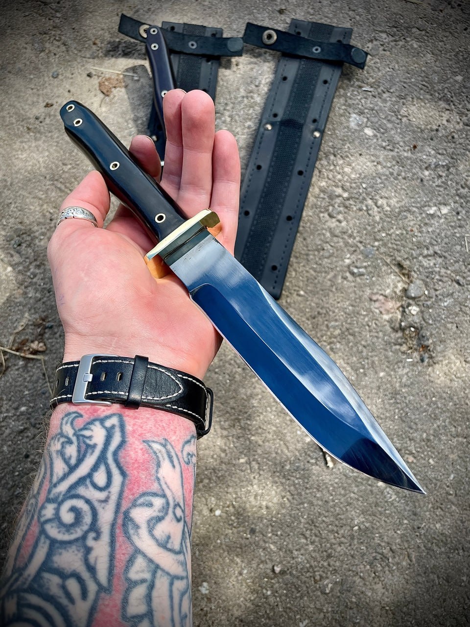 Мужская рука с татуировкой держит тактический нож Warrior Bowie Black Series с черным клинком на фоне бетона.