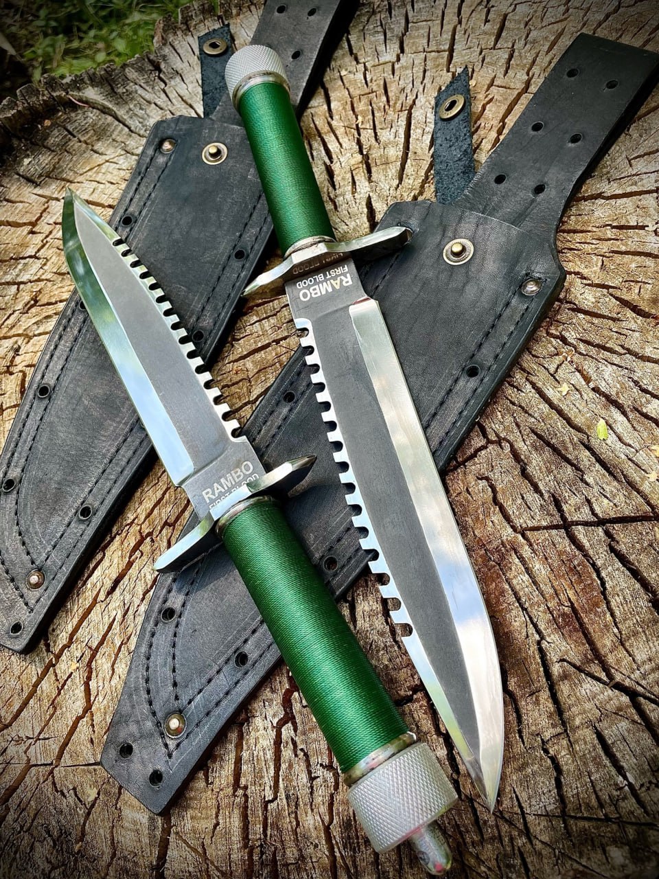 Крупный план двух ножей «Рэмбо: Первая кровь, часть 1» с гравировкой RAMBO FIRST BLOOD на клинке, зелёными рукоятями и пилой по всей длине обуха на кожаных ножнах