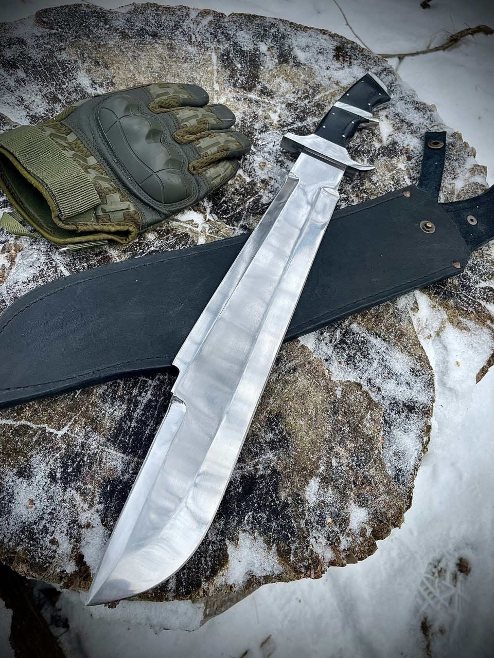 Мачете Билли Сола с широким полированным клинком и чёрными накладками G10 лежит на кожаных ножнах на заснеженном камне рядом с тактической перчаткой