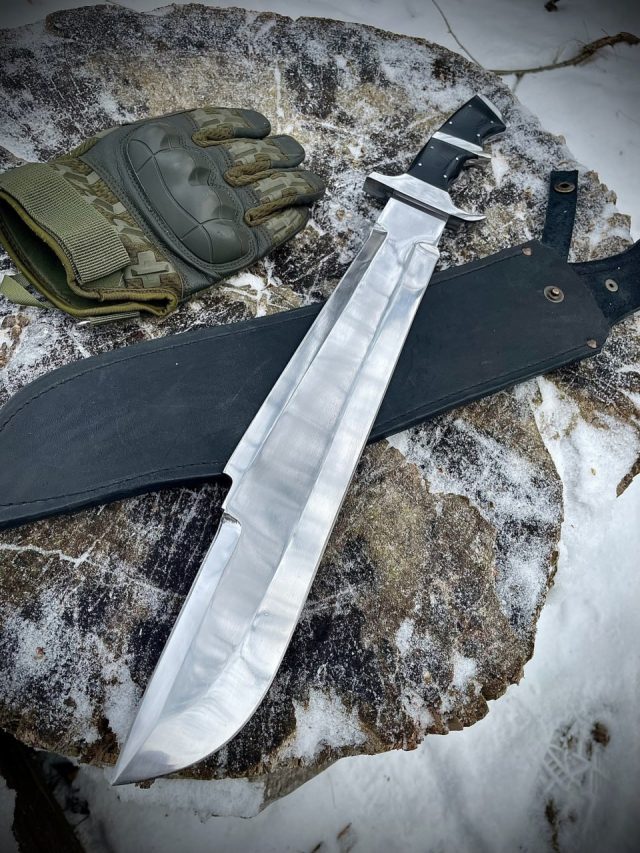 Мачете Билли Сола с широким полированным клинком и чёрными накладками G10 лежит на кожаных ножнах на заснеженном камне рядом с тактической перчаткой